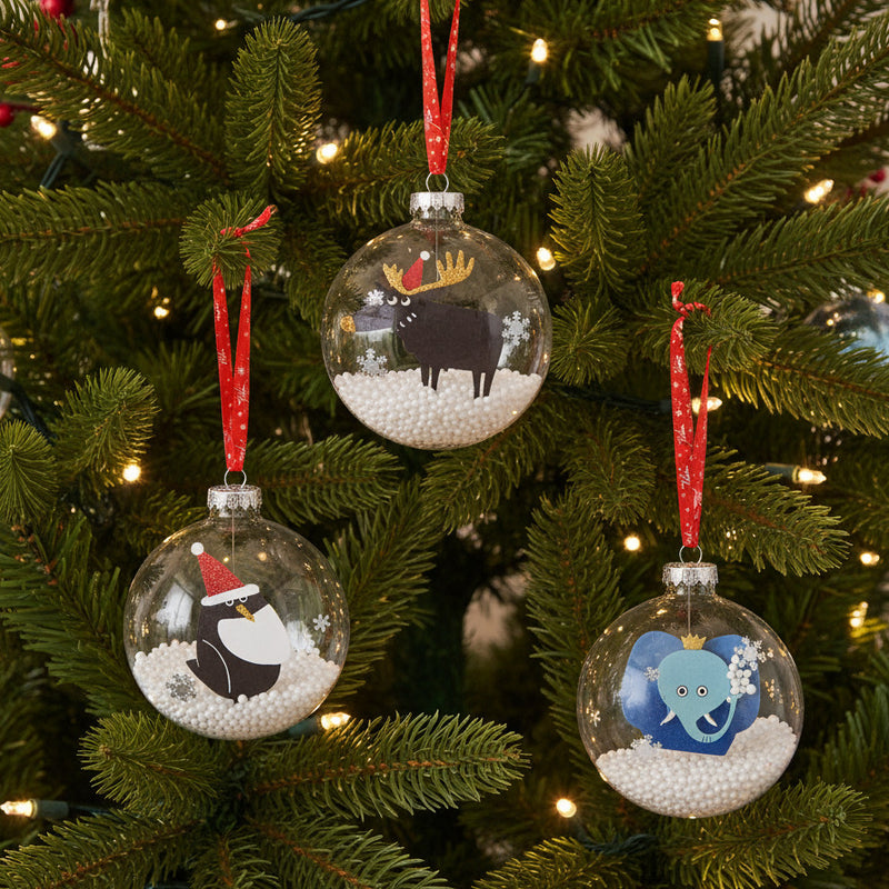 Three Christmas ornaments with penguin, moose, and elephant designs hanging in lighted tree.