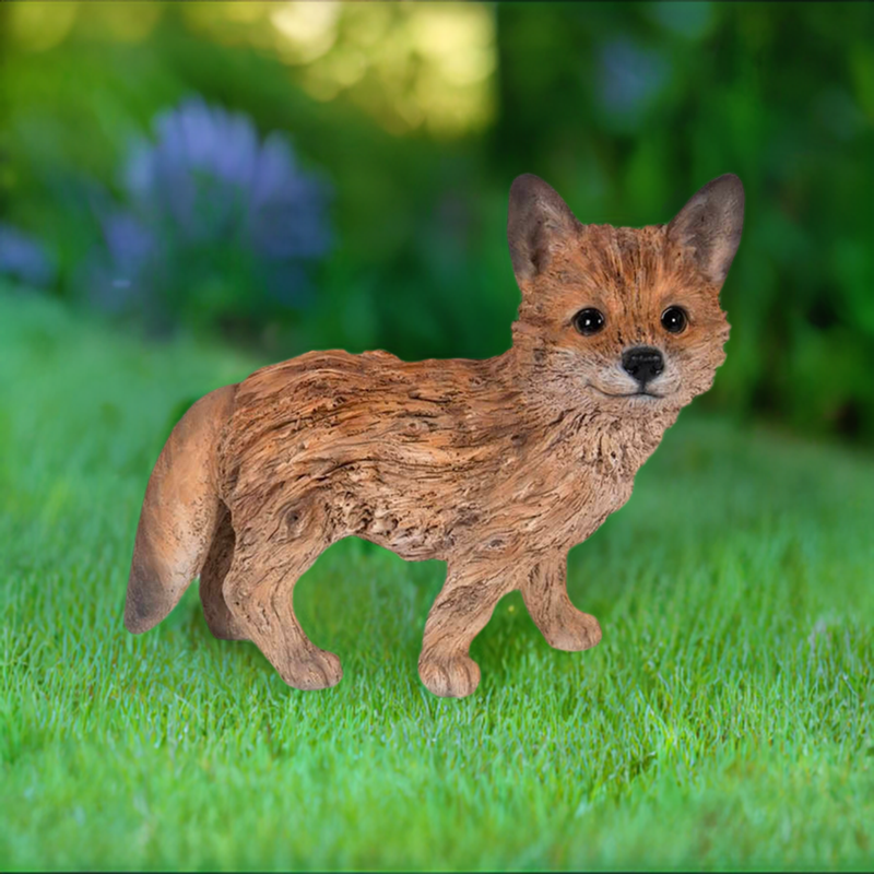 driftwood look baby fox figurine standing on grass with flower garden and trees in background