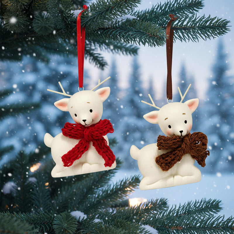 Two Department 56 porcelain reindeer ornaments with red and brown scarves hanging in tree with winter scene in background