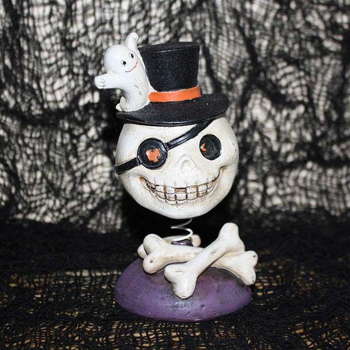 halloween skull with eye patch in top hat with ghost bobble head figurine front view showing crossed bones below head with spider web background